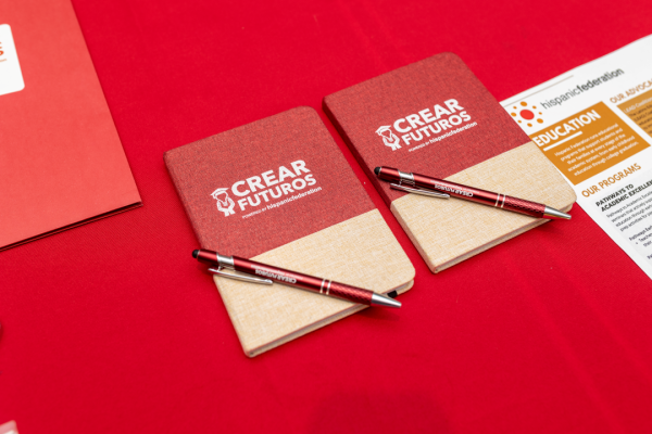Two red and white notebooks with CREAR Futuros printed on them sit on a red table with a pen on top of each book.